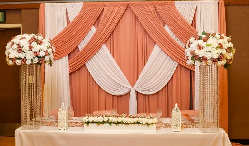 Head table before a blush and ivory draped backdrop, flanked by tall white and dusty-rose rose arrangements on crystal stands