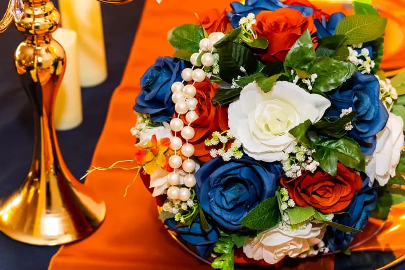 Bouquet of navy, orange and ivory roses wrapped in pearls on an orange satin runner beside a gold candelabra