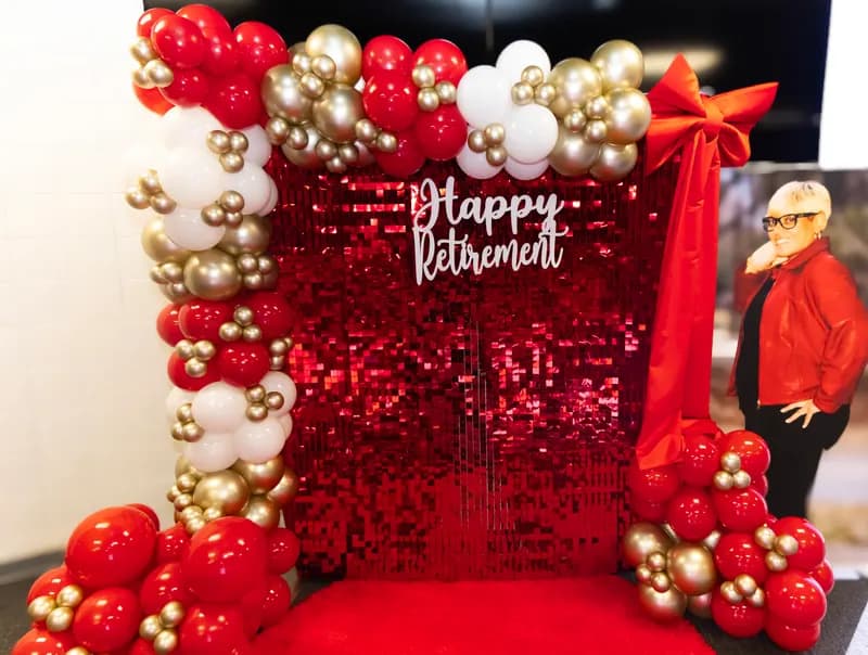 Red sequin 'Happy Retirement' backdrop framed by red, white and gold balloon garland with a red bow